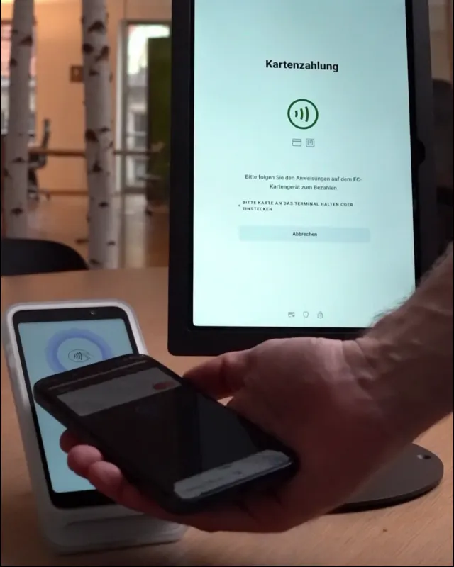 Ein Handy wird kontaktlos an das Kartenterminal gehalten, das Tablet zeigt „Kartenzahlung“.