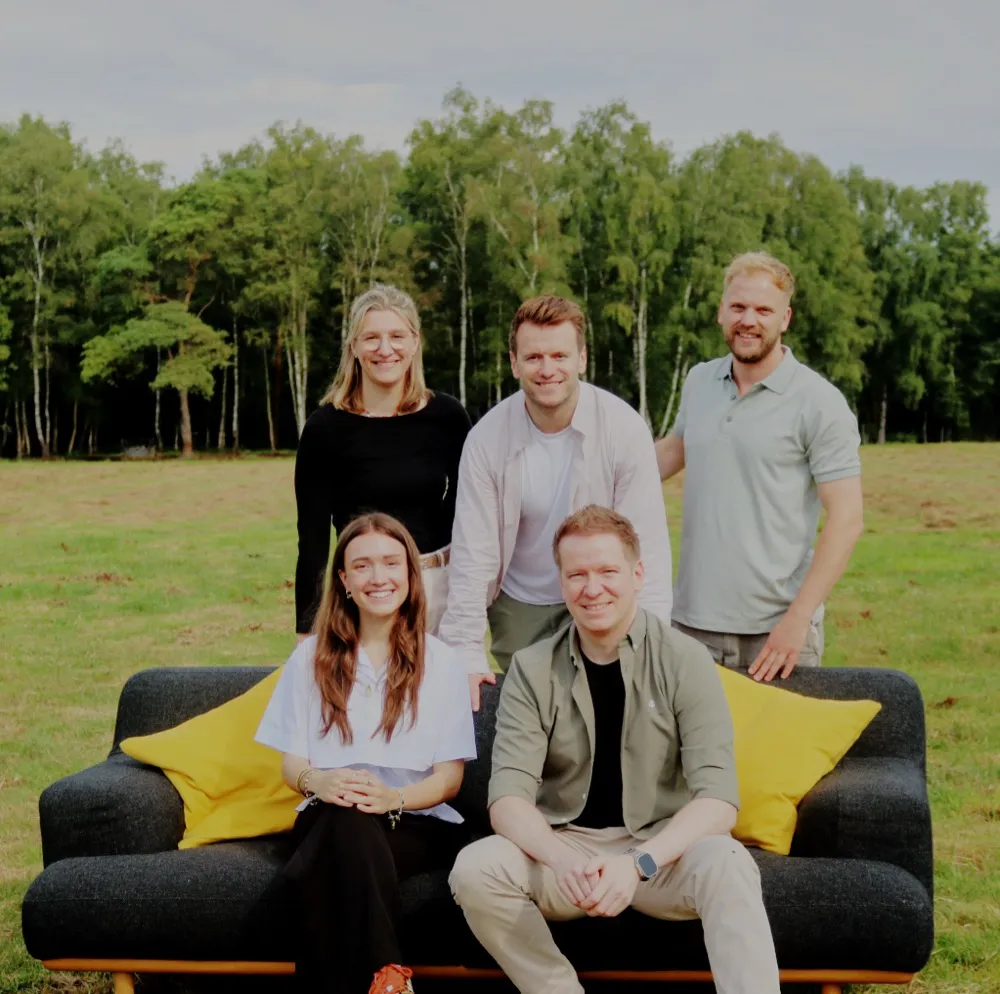 Das Friedhold-Team auf einer Wiese: fünf Personen um ein dunkles Sofa mit gelben Kissen.