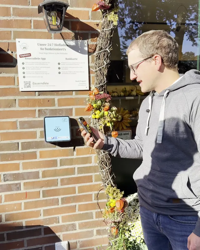 Sven Reuter hält sein Handy an das Kartenlesegerät neben der Ladentür, darüber das Schild „Unser 24/7 Hofladen – So funktioniert's“.
